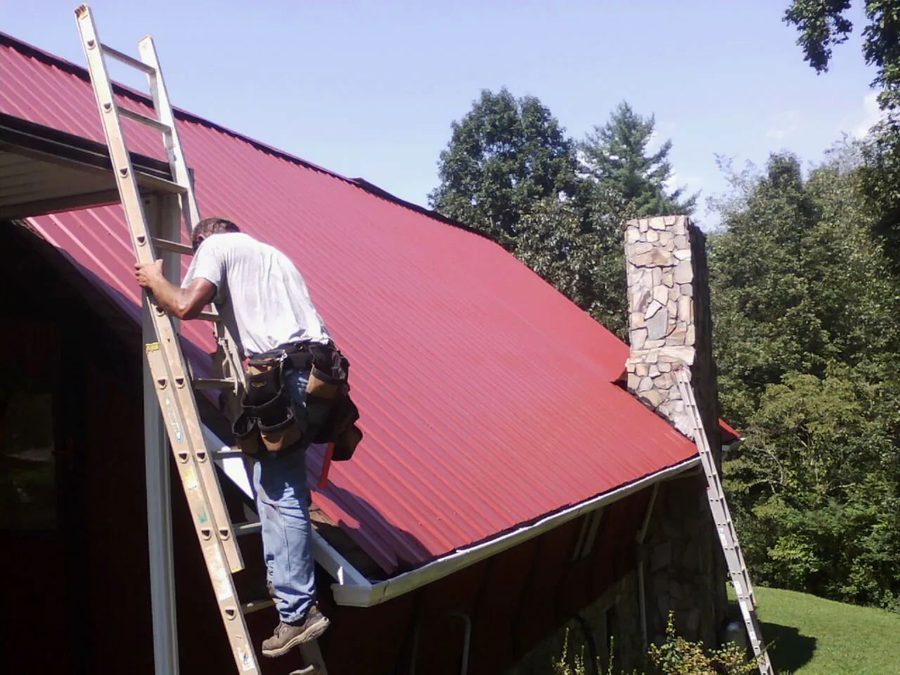 Roofer on a ladder beside a red metal roof and stone chimney — Soffit & Fascia Repair by Roofing Brothers in Pleasanton